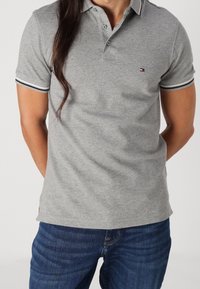 Šedé polo tričko vyrobené z bavlny s texturovaným povrchem. Obsahuje límec, dva knoflíky a námořnické pruhované akcenty na rukávech.