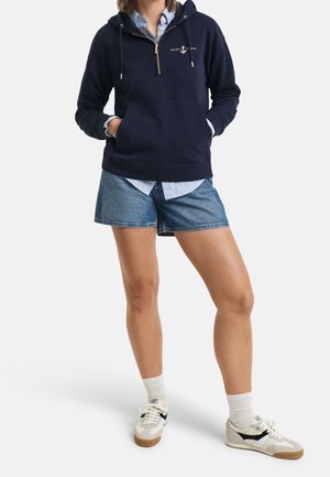 Persona con sudadera con cremallera corta azul marino, camisa de rayas debajo, pantalones cortos de mezclilla, calcetas blancas y zapatillas blancas con detalles en negro y beige.