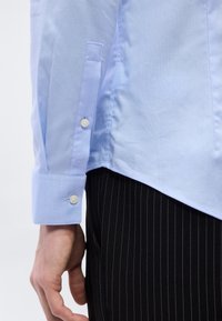 Μανσέτα γαλάζιου πουκαμίσου με κουμπιά και διακριτικό λογότυπο "cK", φορεμένη με μαύρο παντελόνι με ρίγες, χέρι χαλαρό στο πλάι.