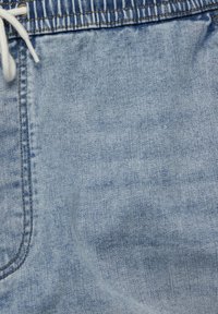 Tissu en denim bleu clair avec des coutures visibles, une taille élastiquée et un cordon blanc noué sur le côté gauche.