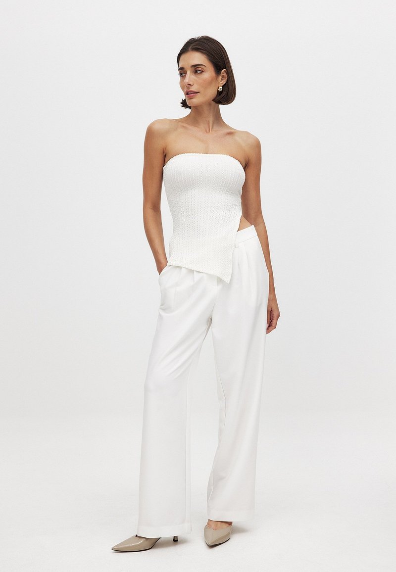 Witte strapless top met geribbelde textuur en asymmetrische zoom, gecombineerd met hoogwaardige, vloeiende witte broeken en puntige beige hakken.