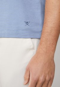 Läheltä kuvattu käsi, joka on vaaleansinisen raidallisen paidan vieressä. Paidassa on pieni brodattu logo ja valkoinen tasku.