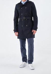 Marinblå trenchcoat med dubbelknäppta knappar, matchande bälte och axelklaffar. Jordad med en ljusblå skjorta och blå byxor, samt vita sneakers.