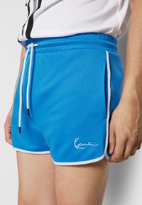 Blå treningsshorts med hvit snøring, sidelommer, hvit kant og brodert logo på den nedre kanten. Laget av et glatt stoff.
