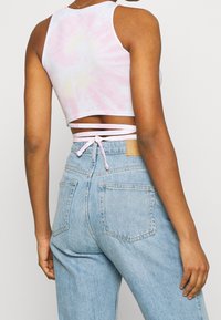 Top corto sin mangas en tonos pastel tie-dye con matices en rosa y amarillo, con un panel trasero recortado y detalles de encaje entrecruzado. Combinado con jeans de mezclilla azul claro.