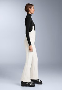 Fehér bordázott kantáros nadrág szárnyas szárakkal, fekete hosszú ujjú felsőre vetve, vastag fekete bakancsokkal, ezüst díszítéssel.