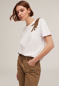 Camiseta blanca de algodón con un adorno decorativo de cuentas marrones en el hombro izquierdo. Combinada con pantalones beiges.