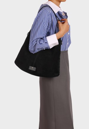 Persona con camicia a righe blu e gonna grigia che tiene una grande borsa nera a tracolla sulla spalla destra su sfondo semplice.