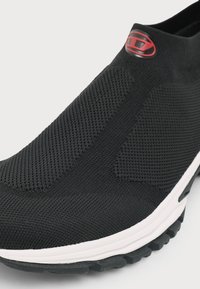 Κοντινό ενός μαύρου πλεκτού sneaker slip-on με υφή ύφασμα, λευκή σόλα και ένα μικρό κόκκινο λογότυπο κοντά στον αστράγαλο.