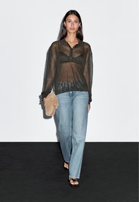 Durchsichtige braune Langarmbluse mit Kragen, kombiniert mit hellblauen Jeans und einer beigen Tasche. Das Model trägt schwarze Sandalen, die Haare sind glatt.