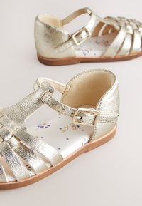 Goldfarbene Metallsandalen mit einem geflochtenen Obermaterial, verstellbarem Riemen und einer Innensohle mit Blumenmuster. Flache Sohle mit brauner Gummilaufsohle.