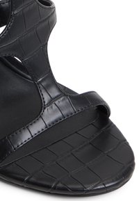Sandal con tacco nero realizzato in pelle texturizzata con motivo a coccodrillo. Presenta un design a ritaglio e una cintura larga sulla zona delle dita.