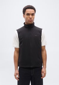 Άνδρας με κοντά σγουρά μαλλιά που φοράει μαύρο γιλέκο με φερμουάρ πάνω από λευκό κοντομάνικο πουκάμισο και σκουρόχρωμο τζιν, στέκεται μπροστά σε ένα απλό φόντο.