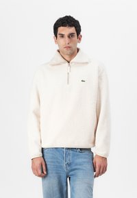 Fleece pullover in light beige with a half-zip neckline, featuring long sleeves and a green logo detail on the chest. Soft texture.