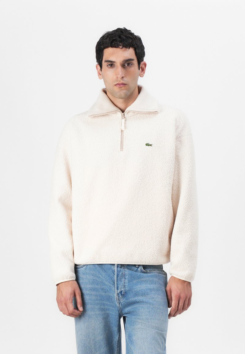 Fleece pullover in light beige with a half-zip neckline, featuring long sleeves and a green logo detail on the chest. Soft texture.