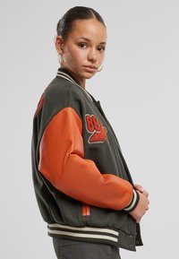 Grønn ull bomberjakke med oransje lærærmer, ribbet mansjetter og to sidelommer. Har et brodert design på brystet.