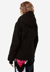 Giacca nera impermeabile con cappuccio, caratterizzata da una texture liscia e un design aderente. Indossata sopra pantaloni a fantasia nei colori rosa, nero e bianco.