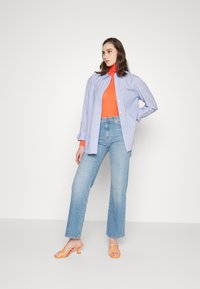 Camisa de manga longa às riscas azuis e brancas sobre uma blusa turtleneck laranja, combinada com jeans de perna reta azul claro e saltos alaranjados de tiras.
