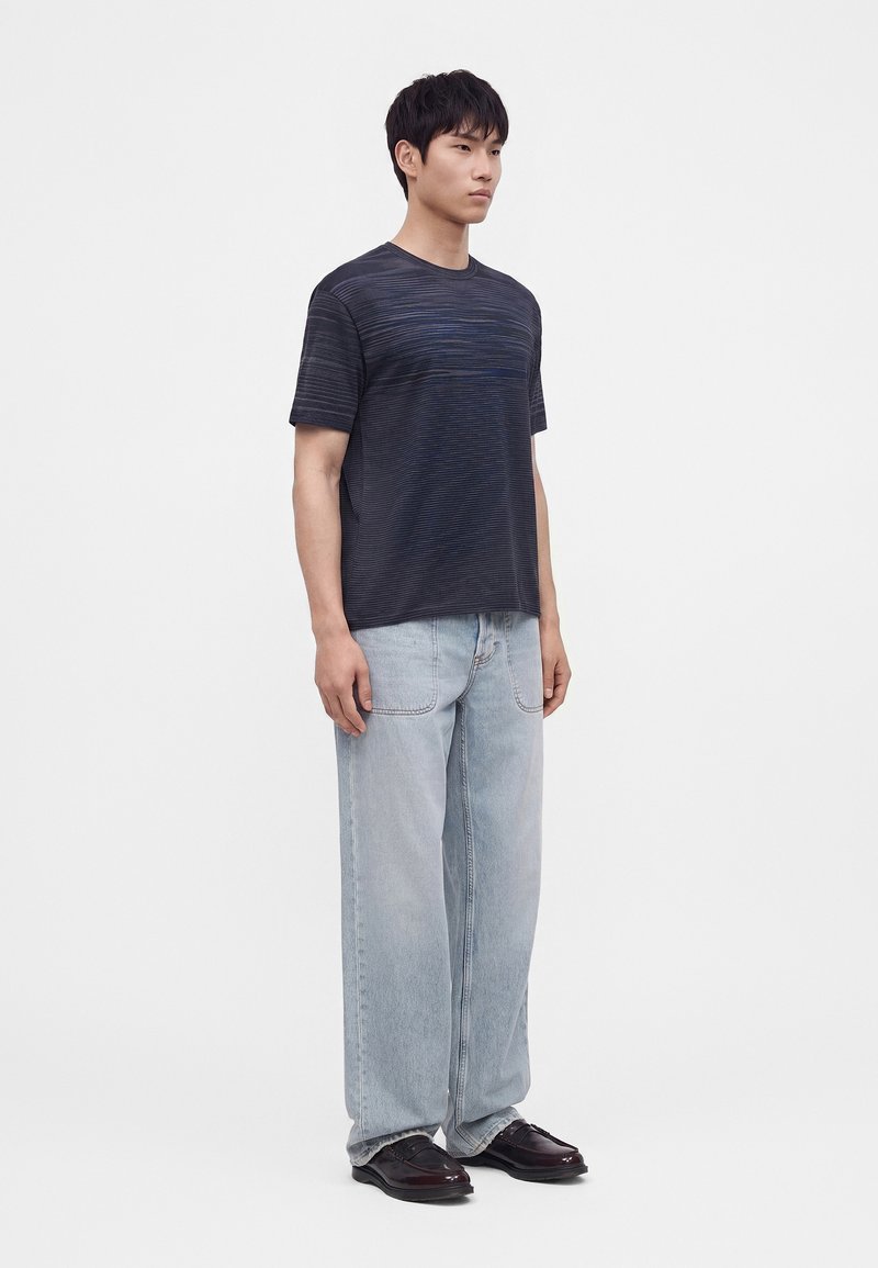 Hombre que lleva una camisa de manga corta a rayas oscuras, vaqueros anchos de color azul claro y zapatos oscuros pulidos, de pie frente a un fondo liso.