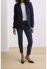 Marinblå blazer med en enkel knapp, matchande skräddarsydda byxor och svarta, spetsiga mule-skor med dekorativa detaljer.
