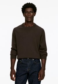 Brauner gestrickter Pullover mit Rundhalsausschnitt und gerippten Bündchen, kombiniert mit dunkelblauen Jeans. Das Material wirkt weich und strukturiert.