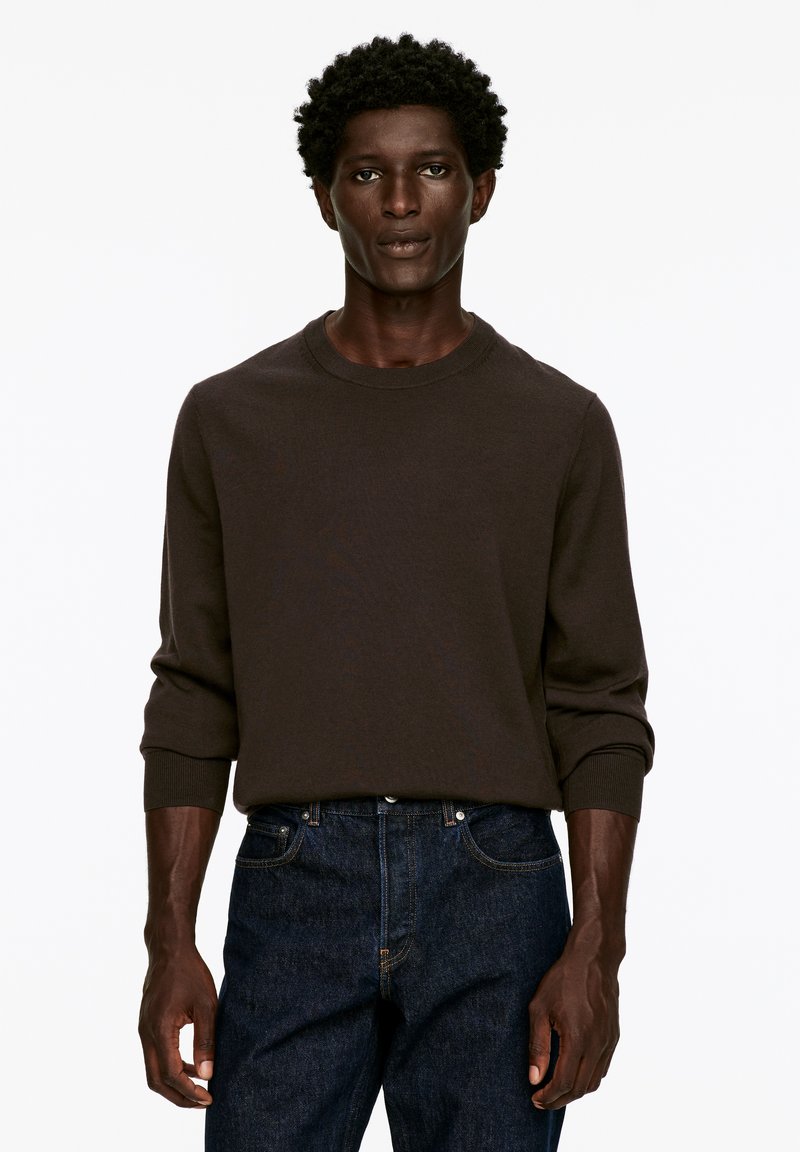 Brauner gestrickter Pullover mit Rundhalsausschnitt und gerippten Bündchen, kombiniert mit dunkelblauen Jeans. Das Material wirkt weich und strukturiert.