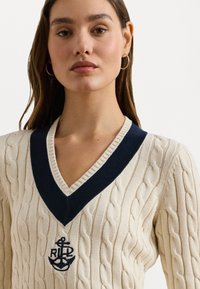 Κρέμα πουλόβερ με πλέξη καλωδίου και βαθύ V-λαιμόκοψη, διακοσμημένο με ναυτικές μπλε λεπτομέρειες, διαθέτοντας ραμμένο λογότυπο άγκυρας στο στήθος. Λείος υφασμάτινος καμβάς.