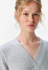 Maglione grigio chiaro a coste con scollatura a V profonda e dettagli di texture subtile. Il tessuto appare morbido e leggermente peloso.