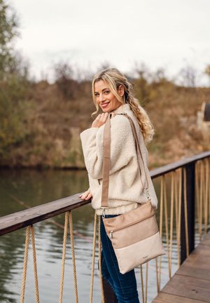 Junge Frau in einer beigen Fleecejacke und blauen Jeans steht auf einer Holzbrücke am Wasser, lächelt und hat eine beigen Umhängetasche über der Schulter.