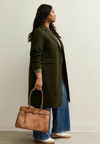 Cappotto lungo verde oliva, jeans in denim a gamba larga, sneakers di colore chiaro. Borsa in suede marrone con dettagli in pelle, portata a mano.