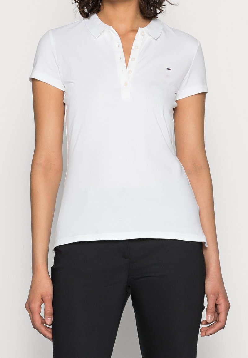 Femme portant un polo blanc à manches courtes avec des boutons et un petit logo sur la poitrine, associé à un pantalon noir.