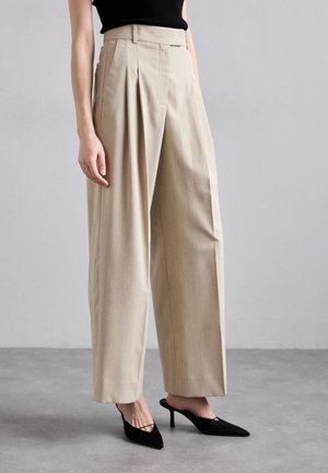 Pantalons beiges taille haute avec plis, en tissu texturé, dotés de jambes larges et d'une coupe décontractée. Portés avec des escarpins noirs à bout pointu.