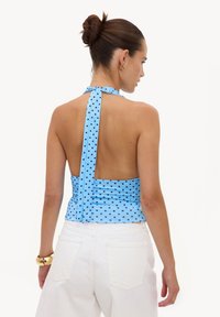 Femme aux cheveux bruns en chignon portant un haut dos nu bleu à pois noirs et un pantalon blanc, vue de dos sur un fond uni.