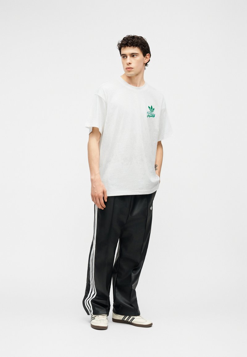 Jeune homme debout portant un t-shirt blanc Adidas, un pantalon de survêtement noir Adidas avec des rayures blanches, et des baskets blanches avec des rayures noires.