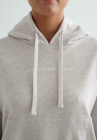Primer plano de una persona que lleva una sudadera ligera gris con un bolsillo frontal y cordones, mostrando texto parcial en el pecho que dice "AIRN* SPORTSWEAR".