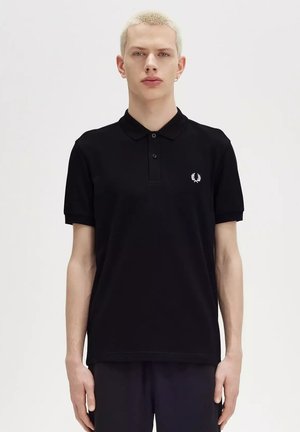 Persona joven con cabello rubio corto que lleva una camiseta polo negra con un pequeño logo blanco de corona de laurel en el pecho, de pie contra un fondo liso.