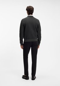 Dunkelgrauer Strickpullover, figurbetontes Design, gerippter Saum und Bündchen, kombiniert mit schwarzen Hosen und schwarzen Schuhen. Einfarbiger weißer Hintergrund.