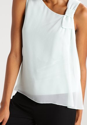 Kvinde iført ærmeløs, lyseblå chiffonbluse med sløjfedetalje på højre skulder og sorte bukser.