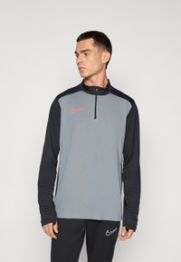 Grijze en zwarte sporttop met lange mouwen en een kwart rits, voorzien van een rood Nike-logo op de borst en zwarte accenten op de mouwen.