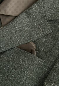 Blazer en tweed vert avec un léger motif à carreaux, comportant un mouchoir de poche marron à motif et une cravate assortie.