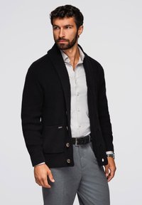 Czarny wełniany sweter z kołnierzem szalowym, z teksturowanej tkaniny, z zapięciem na guziki i dwoma przednimi kieszeniami. Nosi się go na jasnej koszuli.