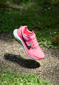 Baskets de sport roses avec une tige en mesh texturé, accents noirs, semelle courbée et logo "ZOOMX" affiché sur le côté. Flottant au-dessus du sol.
