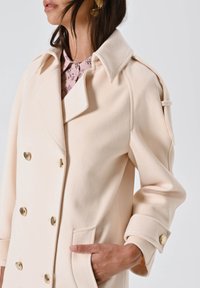 Cappotto beige doppiopetto con bottoni dorati, colletto a pettine e tessuto testurizzato. Presenta tasche laterali e maniche arrotolate.