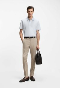 Jasnoniebieska koszulka polo, beżowe spodnie, brązowe mokasyny, czarny pasek, trzyma czarną torbę. Dopasowany krój, krótkie rękawy, subtelny kołnierz.