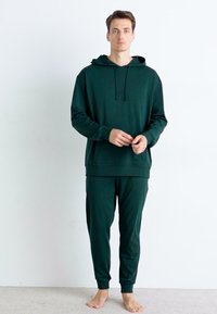 Sweatshirt cu glugă de culoare verde închis și pantalon jogger asortat, confecționat dintr-un material moale, având o croială lejeră și manșete ribbate, pe un fundal simplu.