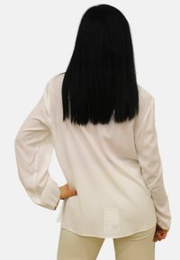 Blusa bianca a maniche lunghe realizzata in tessuto leggero, con orlo curvo, vestibilità ampia e un'etichetta di cura pendente sul retro.