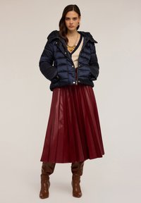 Geacă puffer bleumarin cu glugă, textură matlasată și accesorii aurii, combinată cu o fustă lungă plisată burgundă și cizme maro până la genunchi.