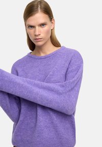 Fioletowy sweter wykonany z dzianiny o miękkiej fakturze, okrągłym dekolcie i obniżonych ramionach. Posiada długie rękawy i swobodny krój.
