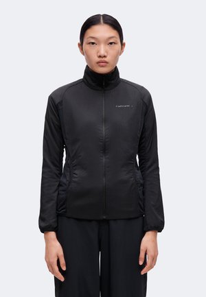 Jeune personne aux cheveux noirs lisses portant une veste noire zippée avec un logo sur la poitrine, debout devant un fond clair uni.