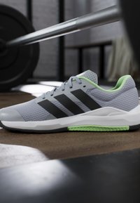 Chaussure de sport gris clair et verte avec des bandes noires placée sur le sol de la salle de sport près d'une barre avec des poids en arrière-plan.